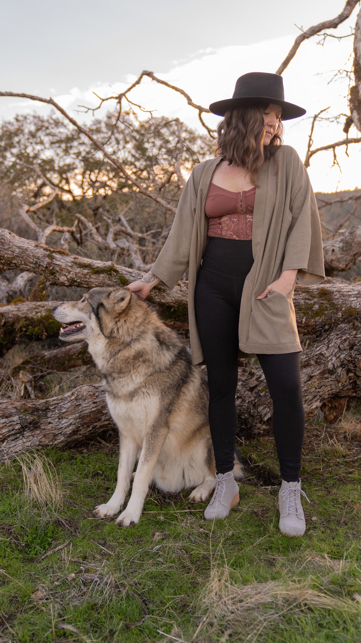 Woman in black hat standing next to large dog wearing black leggings with rose colored top and taupe coverup, trees and sky in the background