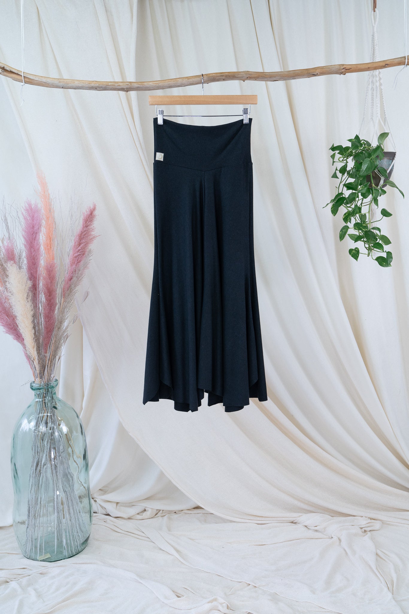 stretchy black, high-waisted, flowing pants on a hanger centered with white fabric backdrop, hanging plant on right and large floral arrangement in glass vase on left