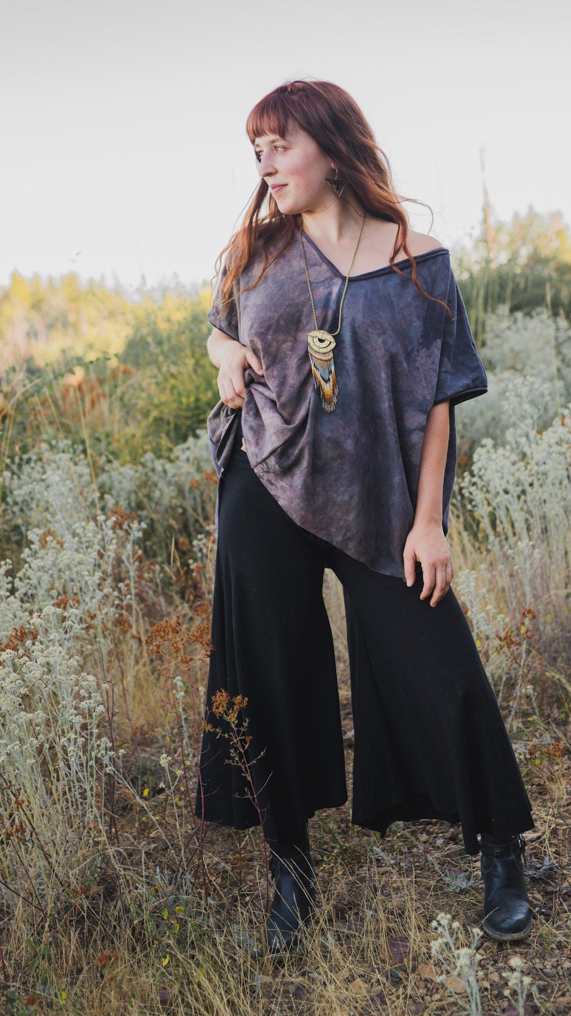 woman with long, red hair and statement necklace standing in a native landscape wearing gray, off-the-shoulder tunic, flowing, black gaucho pants and black boots
