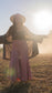 woman with eyes closed and arms out to side standing in light-filled field wearing crop top, robe and stretchy, peachy-pink, high-waisted, flowing pants and a cowboy hat