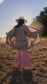 barefoot, sun-lit woman facing away with long brown hair standing in field with trees in background wearing stretchy, peachy-pink, high-waisted, flowing pants and a cowboy hat