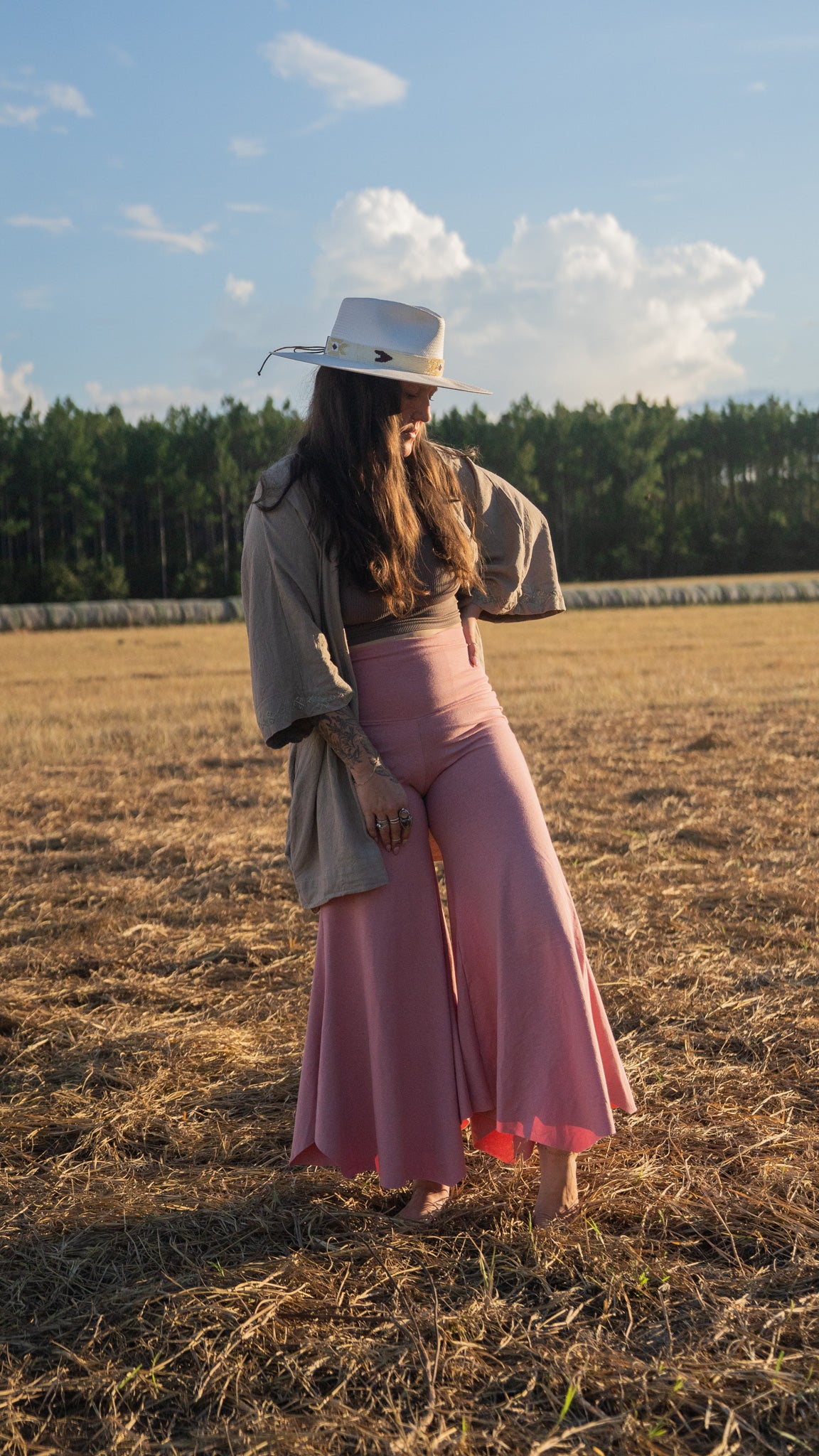 Sun-lit woman with long brown hair standing in field with trees in background wearing stretchy, peachy-pink, high-waisted, flowing pants and a cowboy hat