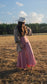 Sun-lit woman with long brown hair standing in field with trees in background wearing stretchy, peachy-pink, high-waisted, flowing pants and a cowboy hat