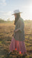 Side-view of barefoot, sun-lit woman with long brown hair standing in field with trees in background wearing stretchy, peachy-pink, high-waisted, flowing pants and a cowboy hat