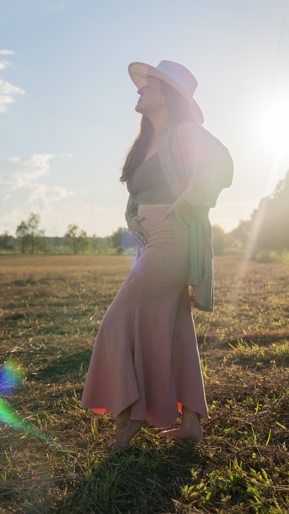 Sun-lit, barefoot woman with eyes closed and hand on belly in field with trees in background wearing stretchy, peachy-pink, high-waisted, flowing pants and a cowboy hat