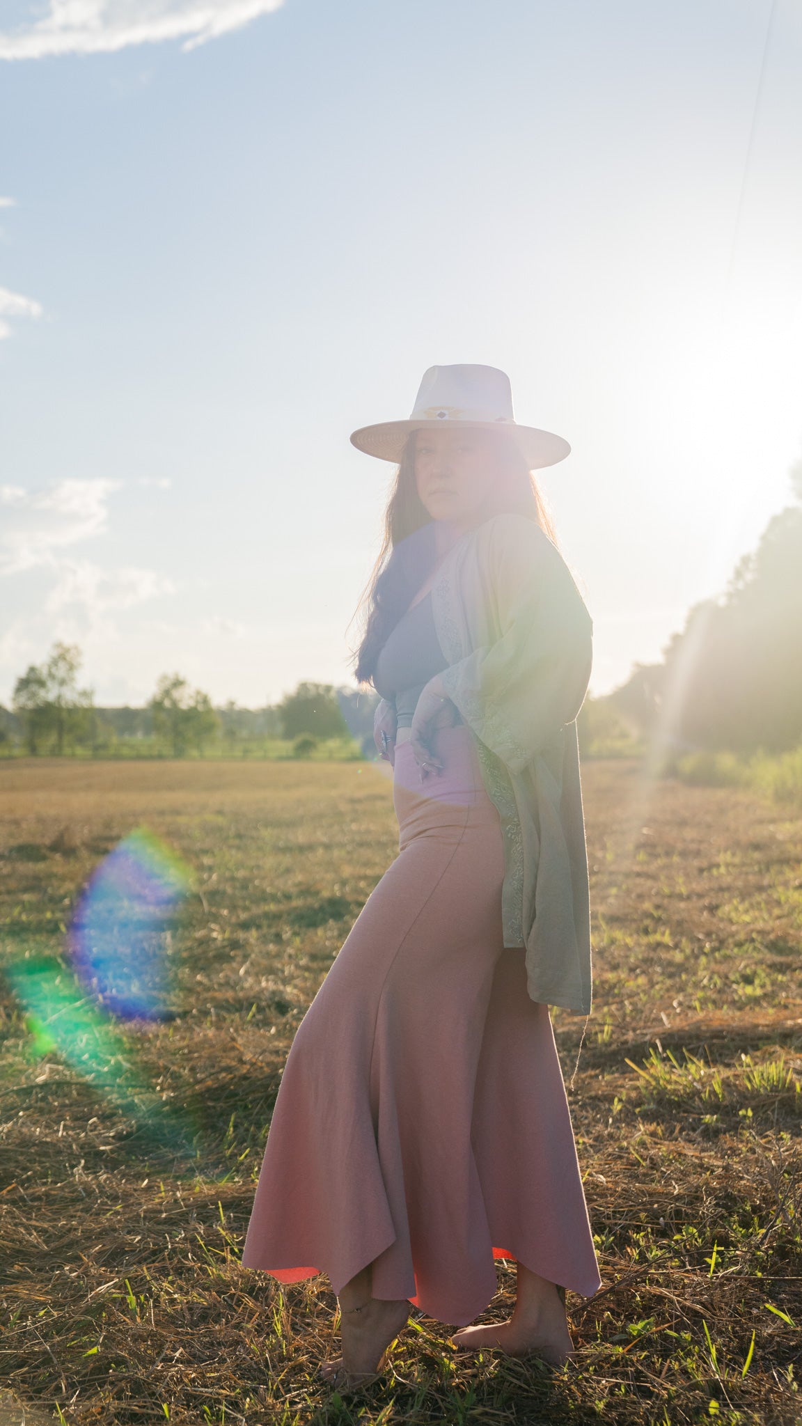 Side-view of barefoot, sun-lit woman with long brown hair standing in field with trees in background wearing stretchy, peachy-pink, high-waisted, flowing pants and a cowboy hat