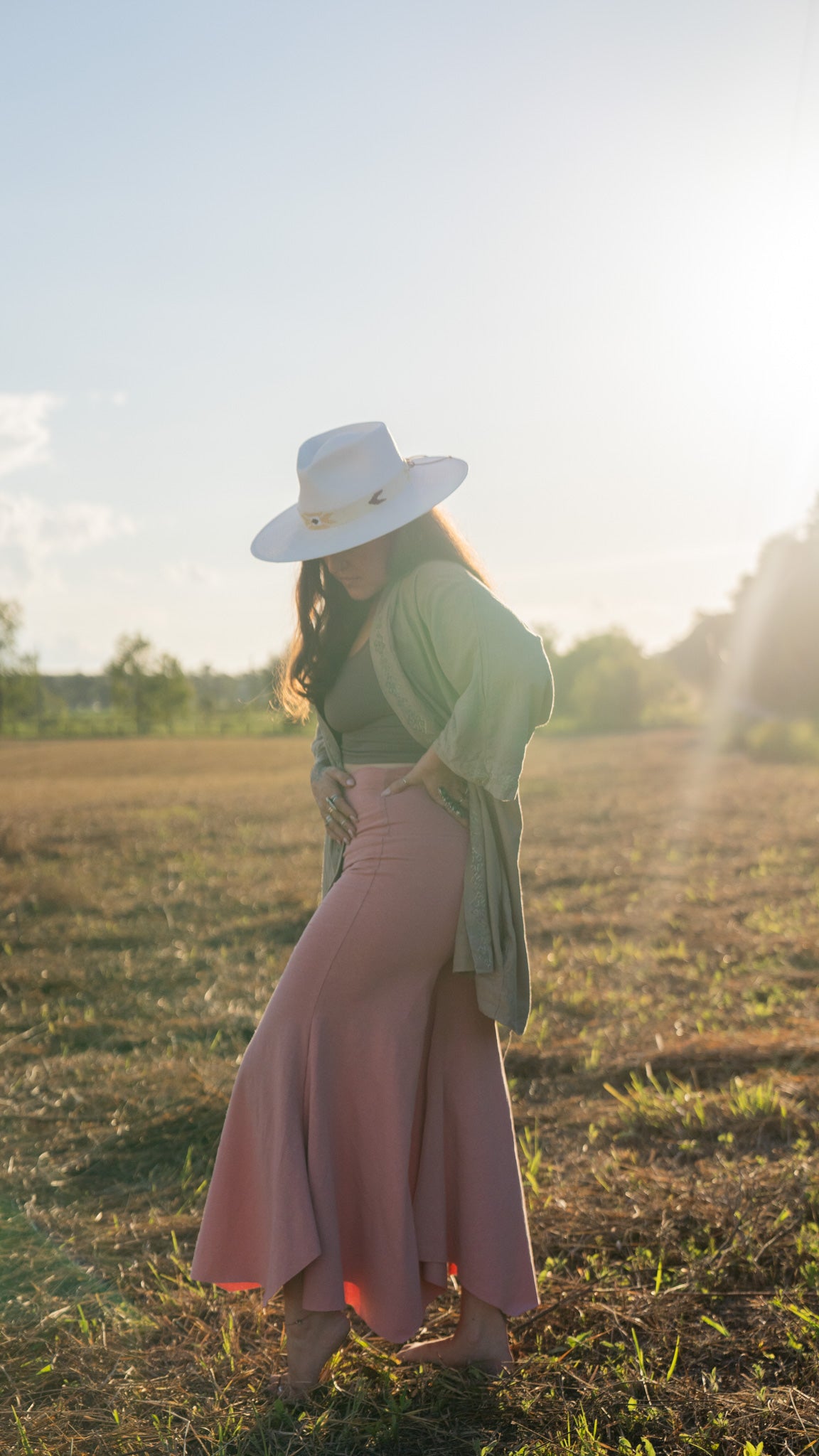 Side-view of barefoot, sun-lit woman with long brown hair standing in field with trees in background wearing stretchy, peachy-pink, high-waisted, flowing pants and a cowboy hat