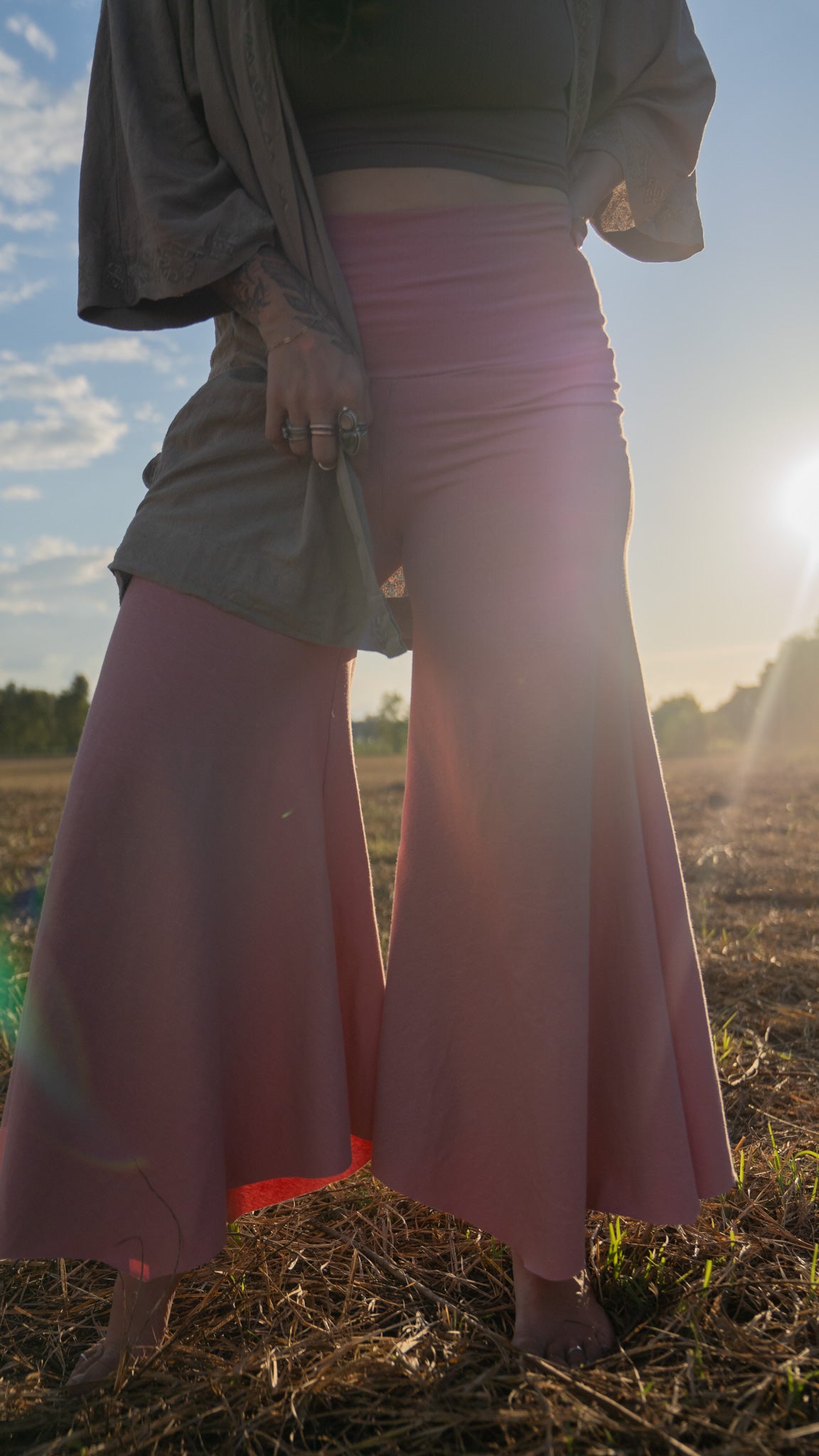 close up of woman standing in sun-lit field with trees in background wearing stretchy, peachy-pink, high-waisted, flowing, gaucho pants