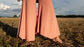 Close up of sun-lit woman standing in field with trees in background wearing stretchy, peachy-pink, high-waisted, flowing pants and an anklet with bare feet