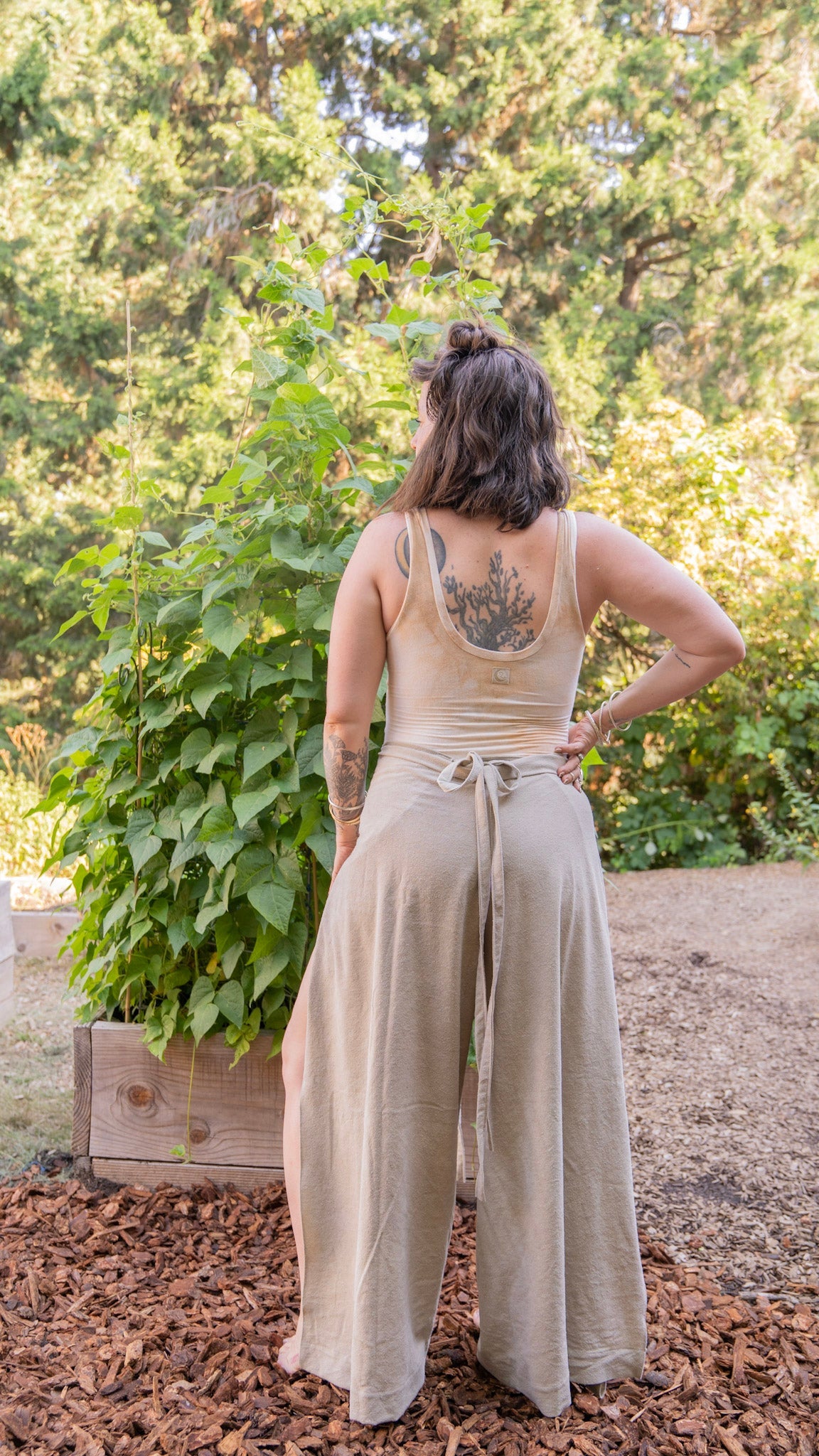 Woman standing outside facing plants in a garden wearing raw silk wrap pants and a yellow leotard