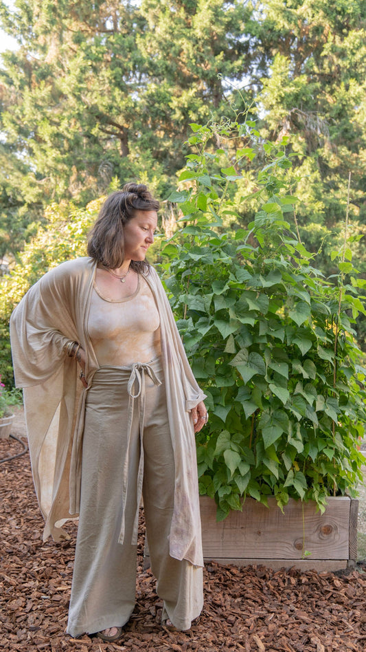 Woman standing in front of plants in a garden wearing raw silk wrap pants, a yellow leotard, and a tan shawl