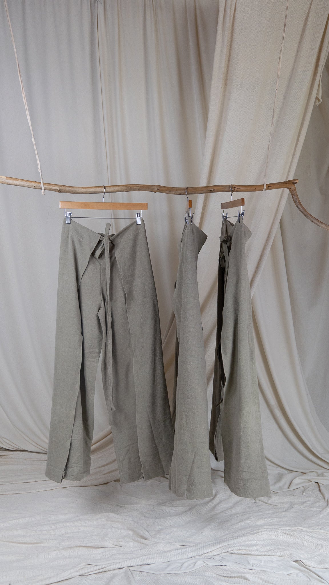 Several pairs of greenish grey raw silk wrap pants hanging from driftwood